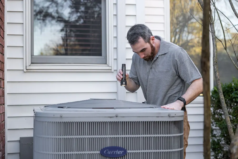 Technician inspecting the top of an outdoor heat pump unit with a flashlight during a service visit